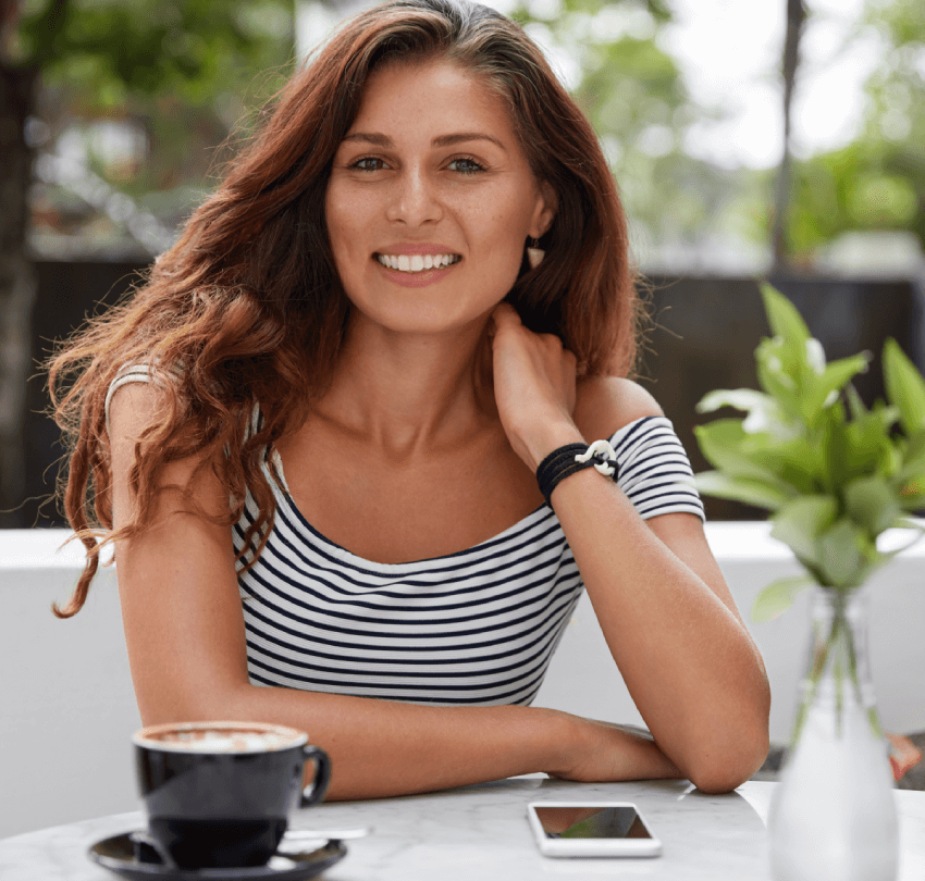 Brunette woman smiling at outdoor café table with coffee, emphasizing wellness and a healthy lifestyle, aligning with natural dental hygiene practices.