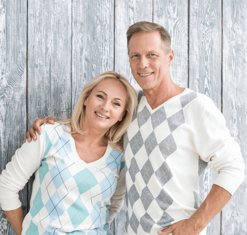 Happy couple smiling together in casual attire, featuring a light blue and white argyle sweater and a gray argyle sweater, against a rustic wooden background, representing the positive outcomes of PRF therapy at Biodental Health and Longevity.