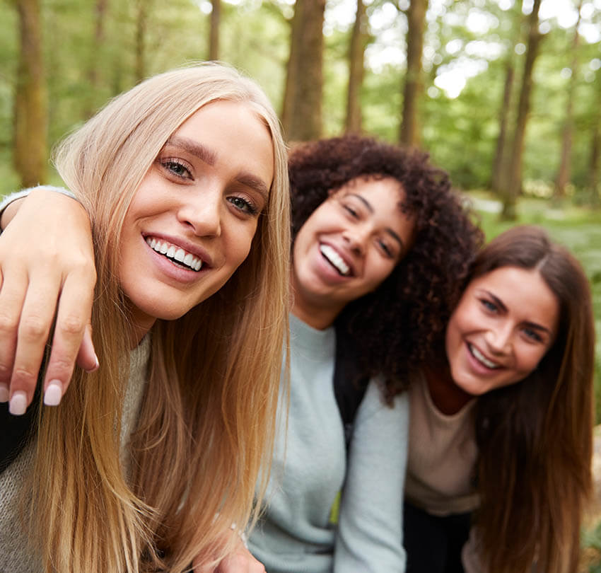 Three women smiling joyfully in a forest setting, showcasing bright, confident smiles, reflecting the theme of summer and cosmetic dentistry enhancements.