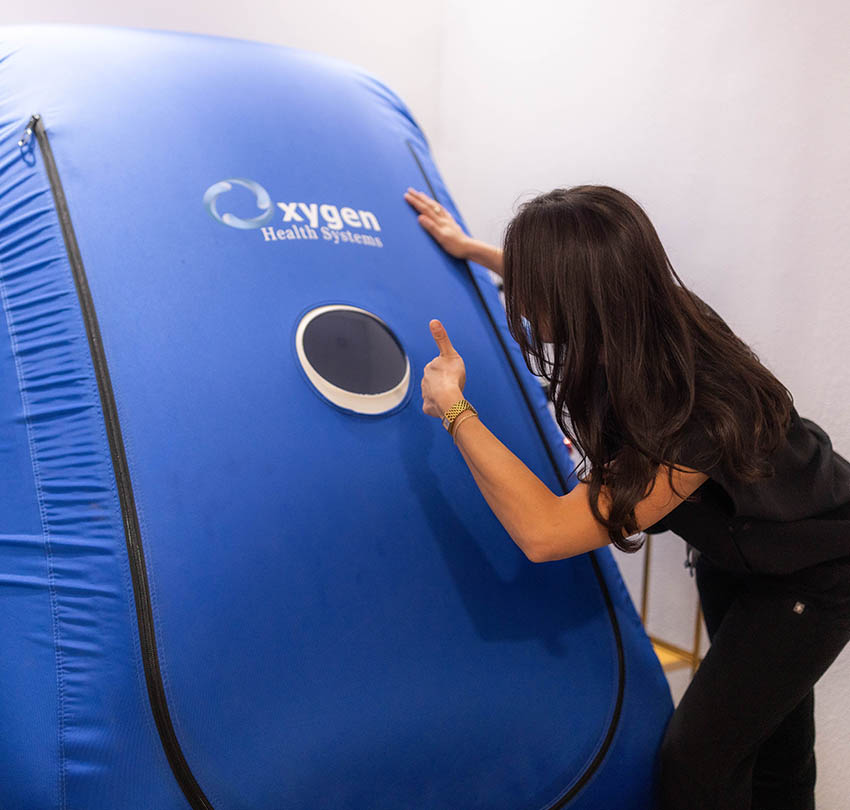Woman demonstrating hyperbaric oxygen therapy equipment with a blue chamber labeled "oxygen Health Systems," promoting health recovery and cognitive clarity.