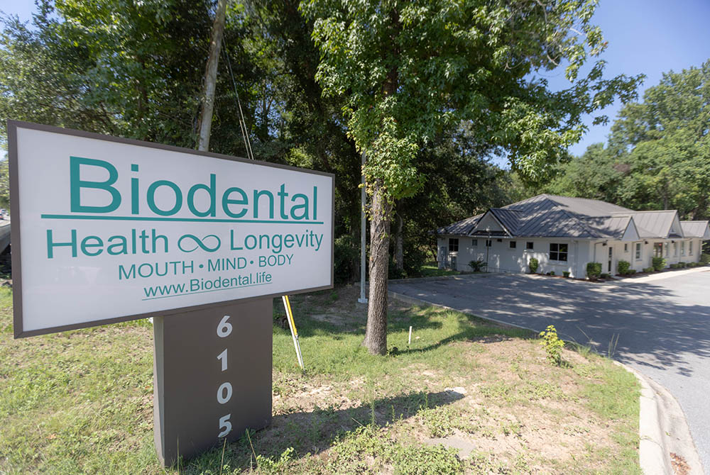 Biodental Health and Longevity office exterior with sign displaying "Biodental Health Longevity MOUTH MIND BODY" and surrounding greenery in Pensacola, FL.